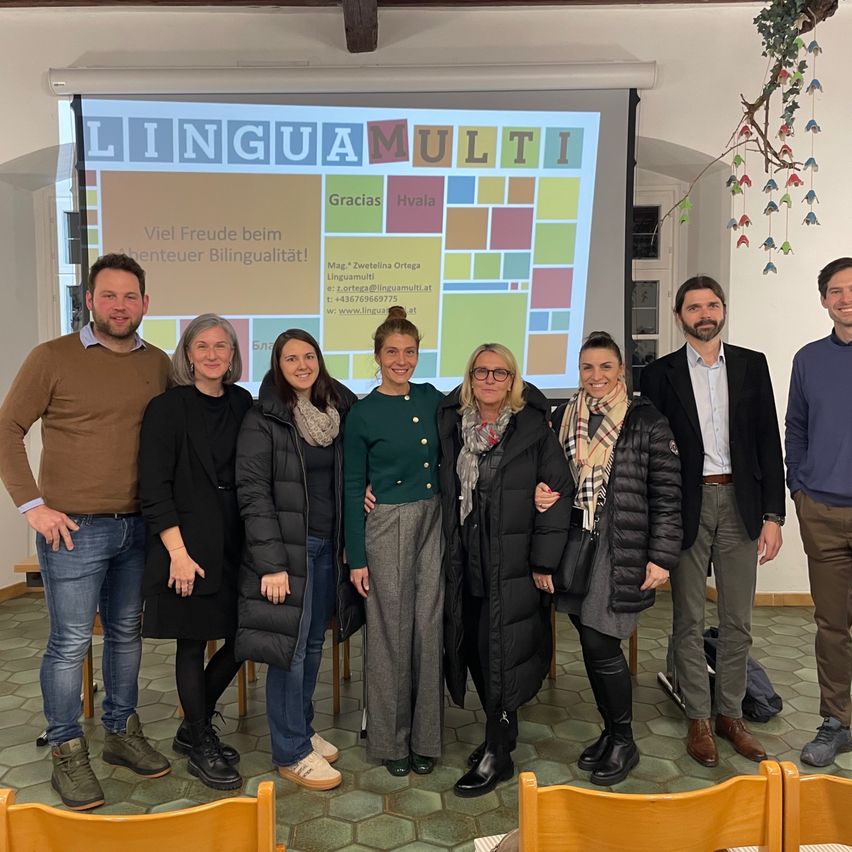 Eine Gruppe von Menschen posiert für ein Foto in einem Raum mit einer Präsentationsleinwand dahinter. Die Leinwand zeigt 'LINGUA MULTI' mit bunten Blöcken und Text.