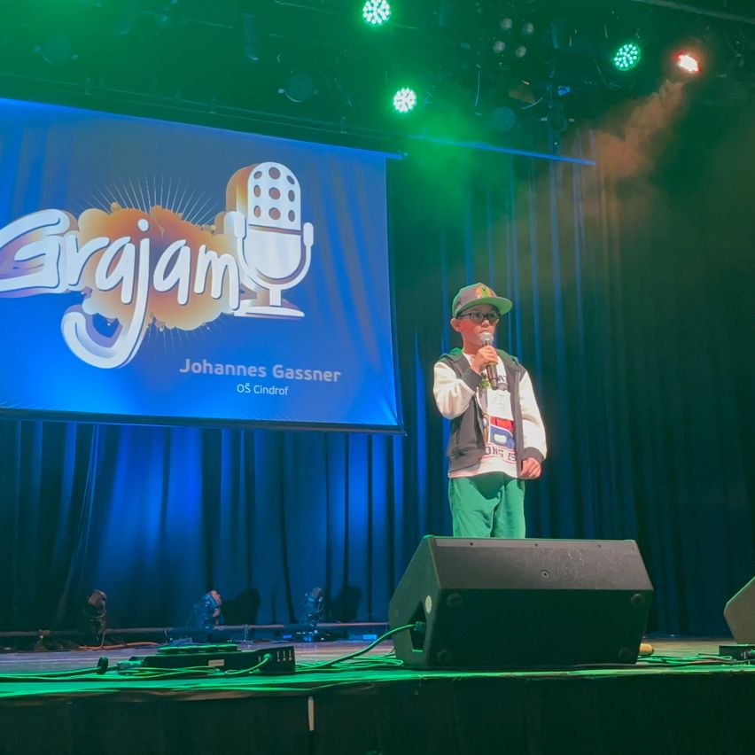 Ein junger Junge steht auf der Bühne und hält ein Mikrofon. Eine große Leinwand hinter ihm zeigt das Wort 'Grgjam' und den Namen 'Johannes Gassner'.