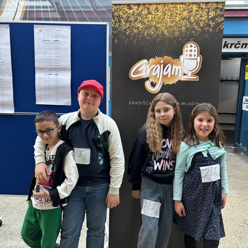 Vier Kinder stehen vor einem schwarzen Poster mit dem Wort 'Grajgam' in Gold und nummerierten Schildern. Sie lächeln für ein Foto. Dahinter zeigt ein blauer Aushang Papiere.