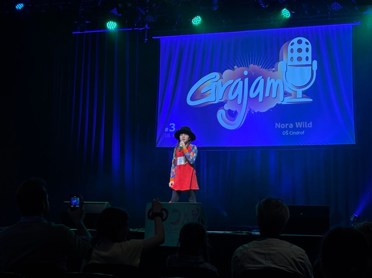 Ein junges Mädchen steht auf der Bühne und hält ein Mikrofon in der Hand. Sie trägt einen Hut und ein farbenfrohes Kleid. Hinter ihr zeigt eine große Leinwand das Wort 'Grajam' mit einem Mikrofon-Symbol. Die Bühne ist mit blauen Lichtern beleuchtet und ein Publikum sieht zu.
