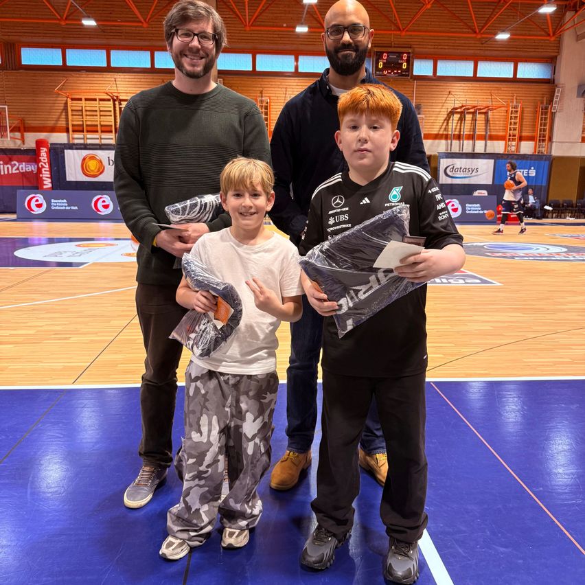 Drei Männer und zwei Jungen auf einem Basketballplatz, mit Preisen in den Händen. Ein Junge in Camouflage-Hosen, der andere in Schwarz. Hinter ihnen ein Basketballkorb und in der Ferne ein Spieler.