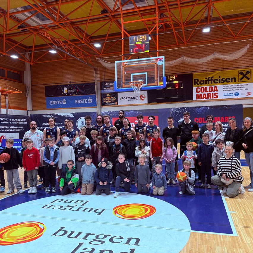 Eine große Gruppe von Menschen, einschließlich Kindern und Erwachsenen, posiert für ein Foto in einer Basketballhalle. Die Halle verfügt über einen großen weißen Kreis mit den Worten 'Purer Energie'. Die Halle wird von 'Burgenland' gesponsert.