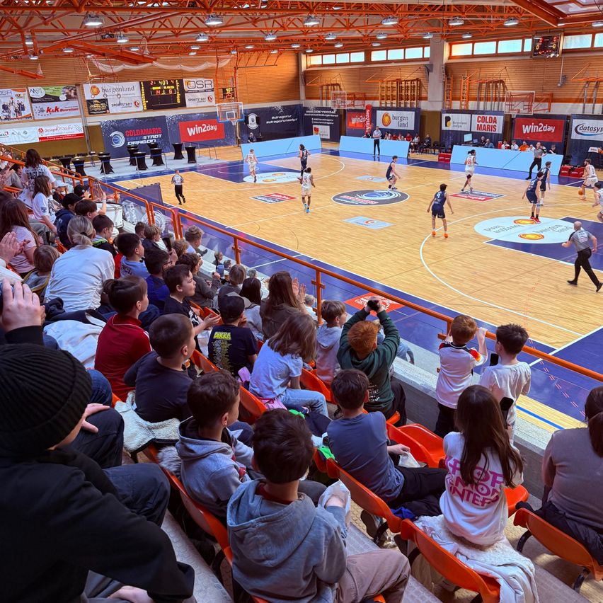 Ein Basketballspiel findet auf einer Indoor-Spielfläche statt. Die Spieler sind aktiv am Spielen, während Zuschauer von den Tribünen zuschauen. Die Spielfläche hat Sponsorenlogos und Werbung an den Wänden.