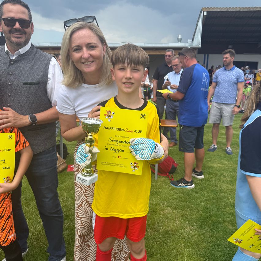 A young boy in a yellow shirt stands with his family, holding a trophy. They are standing on a grassy field, with other families in the background.