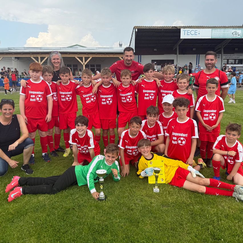 A soccer team in red uniforms poses for a photo, with two players in green and yellow kneeling with trophies. Adults stand behind them, one holding a child. A banner reads 'TECE'.