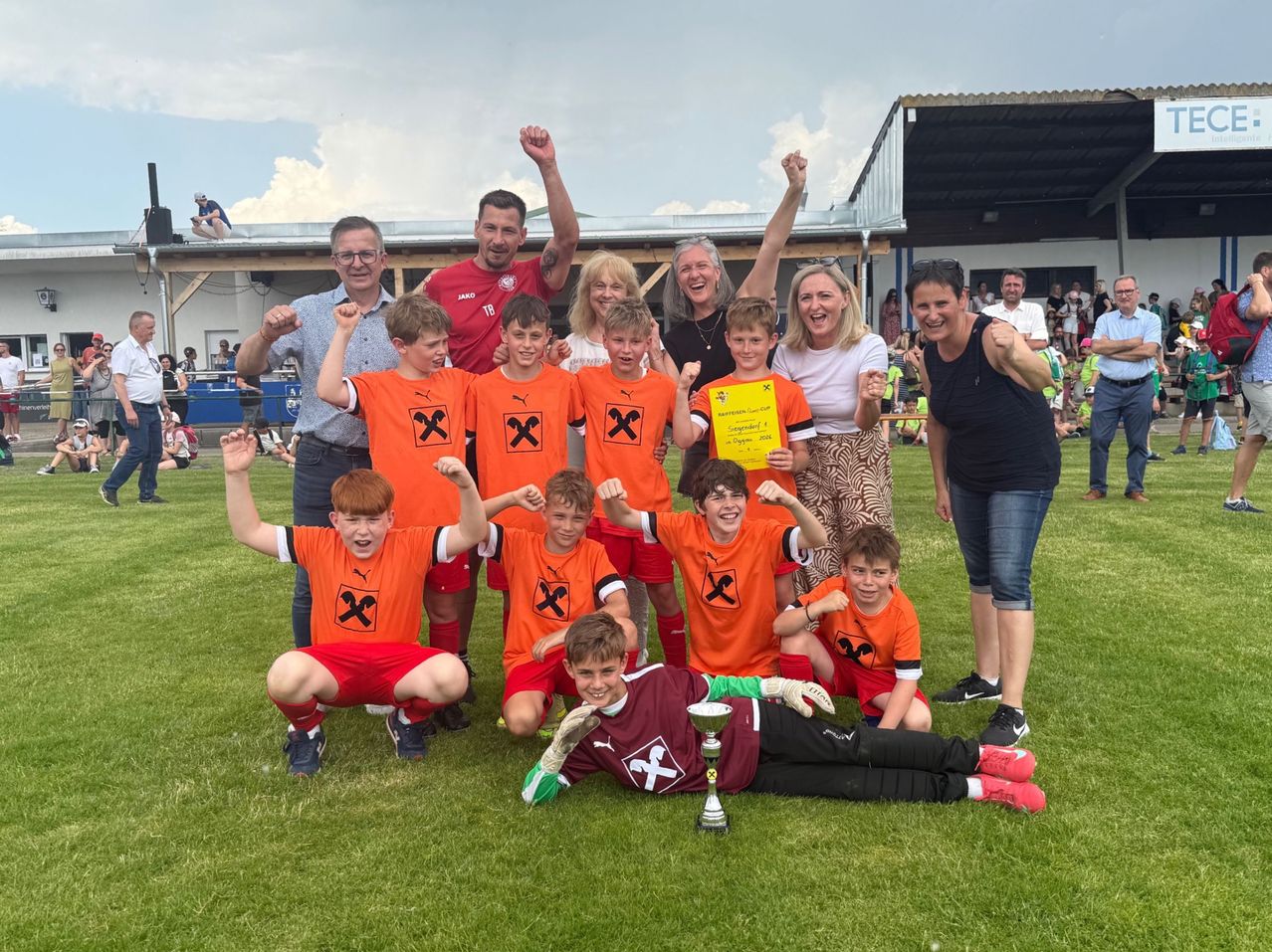 A soccer team in orange jerseys, posing for a photo with adults, holding a trophy, on a sunny field.