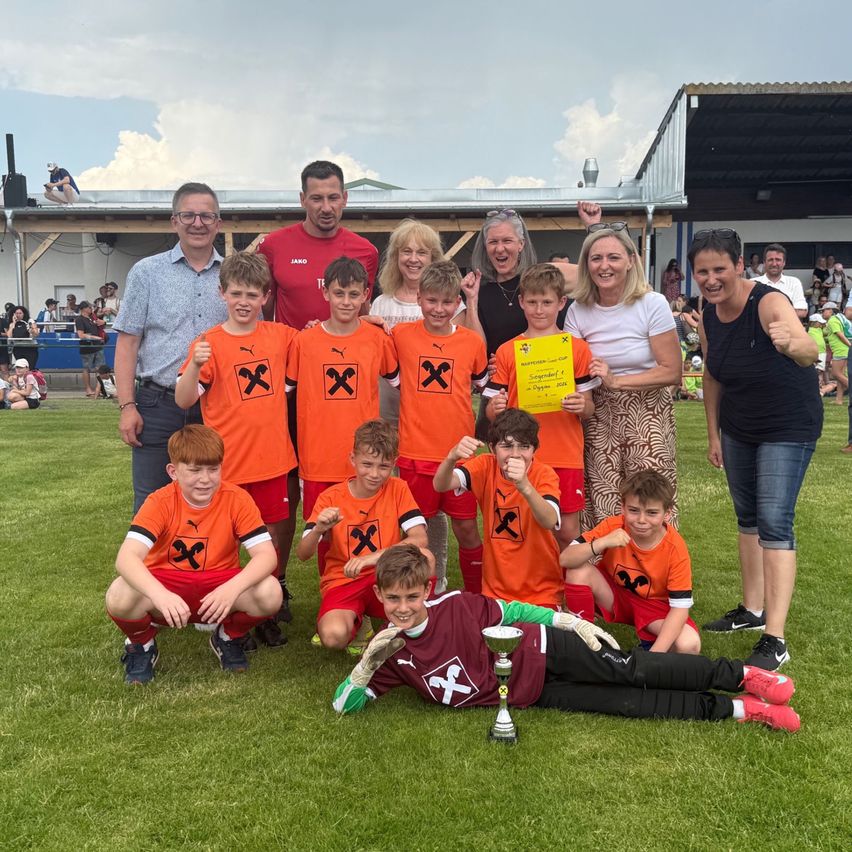 A soccer team of young boys wearing orange jerseys pose with adults for a photo on a field. A boy lies on the ground with a trophy next to him.