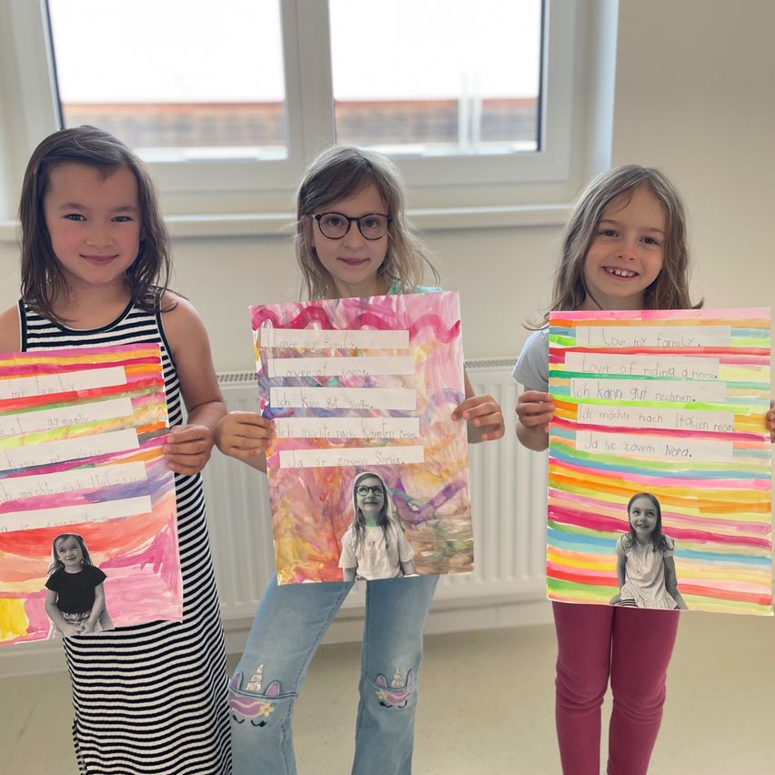 Three young girls hold up colorful artwork with a photo of a girl in the middle. They smile, standing in a room with a window behind them. Each artwork has text and drawings.
