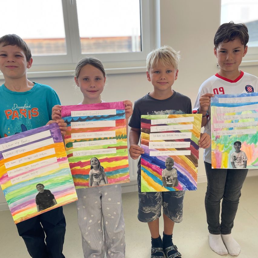 Four children pose for a picture while holding colorful posters with images of themselves and words. They are smiling and seem to be posing for a photo. The posters have a rainbow design and a picture of the children.