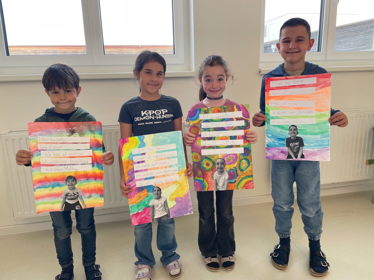 Four children are holding up colorful posters with text and images of people, possibly a family. They are smiling and appear to be in a classroom. The posters have rainbow-like backgrounds.