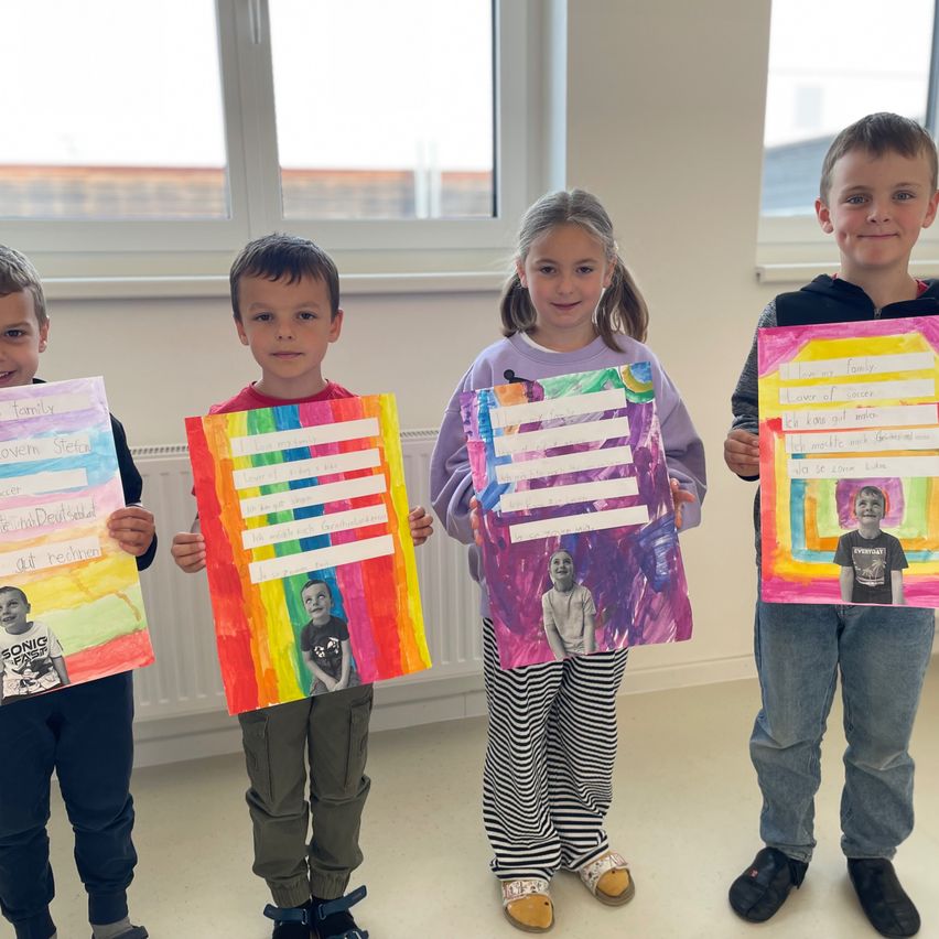 Four children stand together, each holding a colorful artwork. They are smiling and posing for the camera. The artwork includes a picture of a boy and text. The room has large windows and white walls.