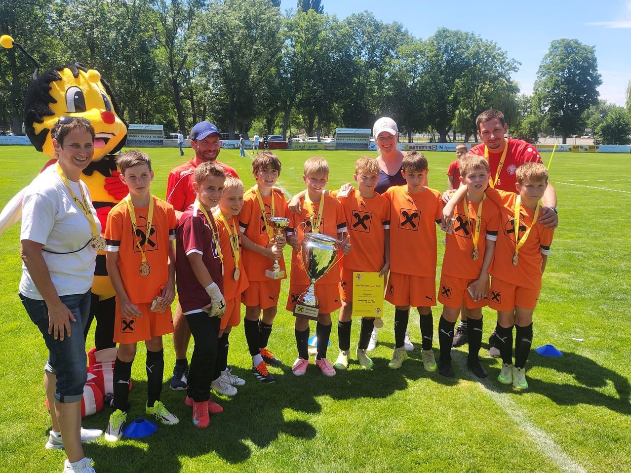 Eine Gruppe junger Jungen in orangefarbenen Uniformen und Medaillen posiert mit einer Trophäe auf einem Feld. Ein Maskottchen und mehrere Erwachsene stehen in der Nähe.