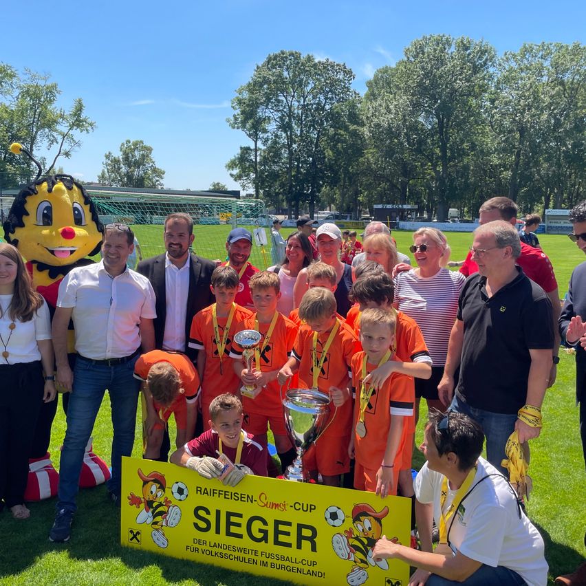 Eine Gruppe junger Fußballer und Erwachsene posiert für ein Foto mit Trophäen und einem Schild, das Sieger sagt. Ein großes Maskottchen steht hinter ihnen.