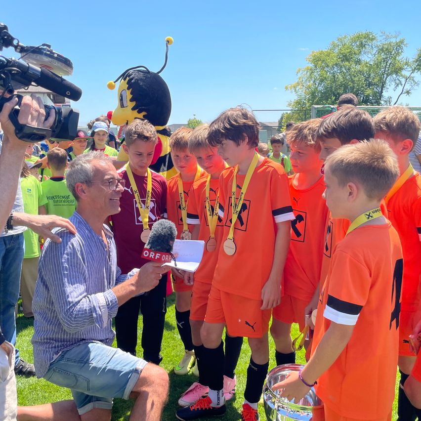 Ein Mann interviewt eine junge Fußballmannschaft mit orangefarbenen Trikots. Sie haben Medaillen und einen Pokal. Ein großes Bienenkostüm ist im Hintergrund.