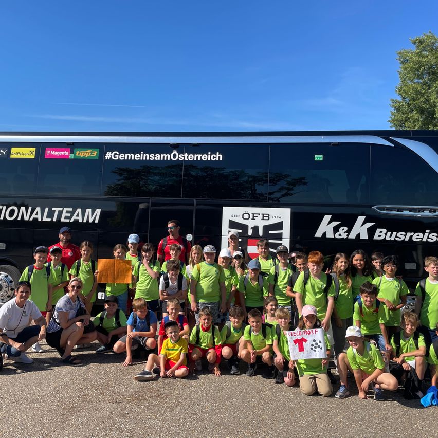 Eine Gruppe von Kindern und Erwachsenen in grünen Shirts posiert für ein Foto vor einem Bus. Der Bus trägt die Aufschriften 'Nationalteam' und '#GemeinsamÖsterreich'.