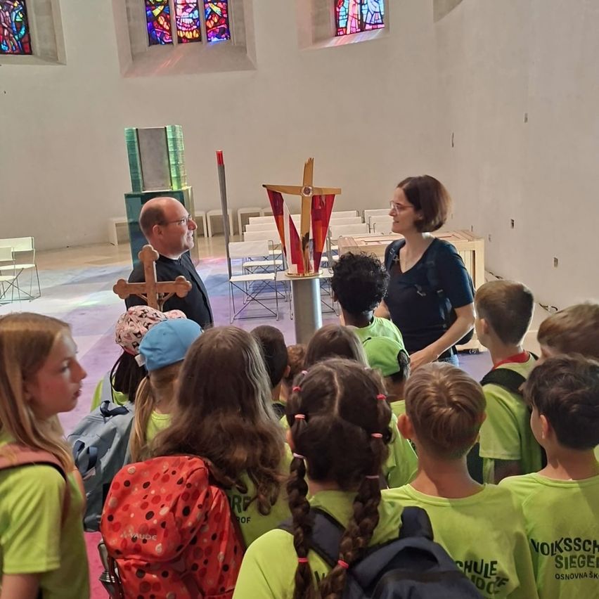 Eine Gruppe von Kindern in grünen Shirts hört einem Priester und einer Frau in einer Kirche zu. Ein Kreuz steht auf dem Altar.