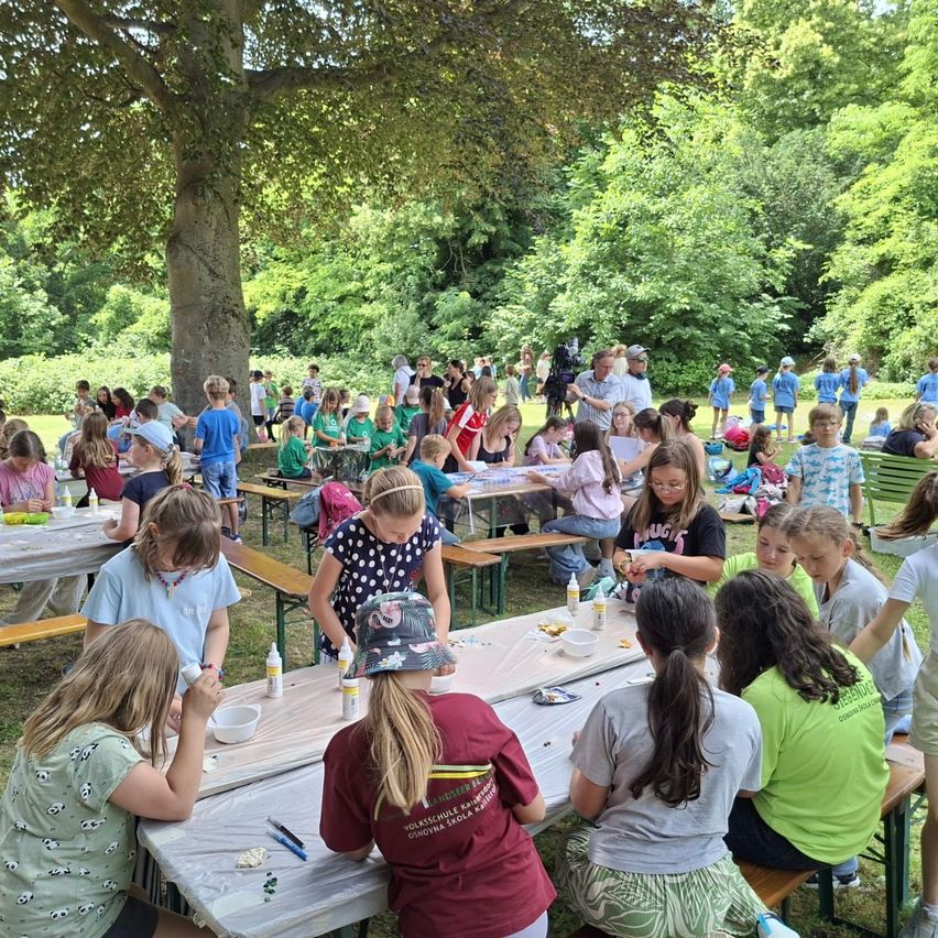 Eine Gruppe von Kindern in grünen Shirts sitzt um Tische in einem Park herum und macht ein Kunstprojekt. Sie sind von Bäumen umgeben.