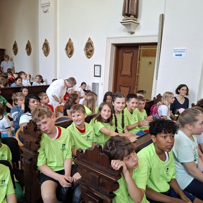 Eine Gruppe von Kindern in grünen Shirts sitzt in Kirchenbänken und schaut einen Mann in Weiß an, mit einer Statue und gerahmten Bildern an der Wand.