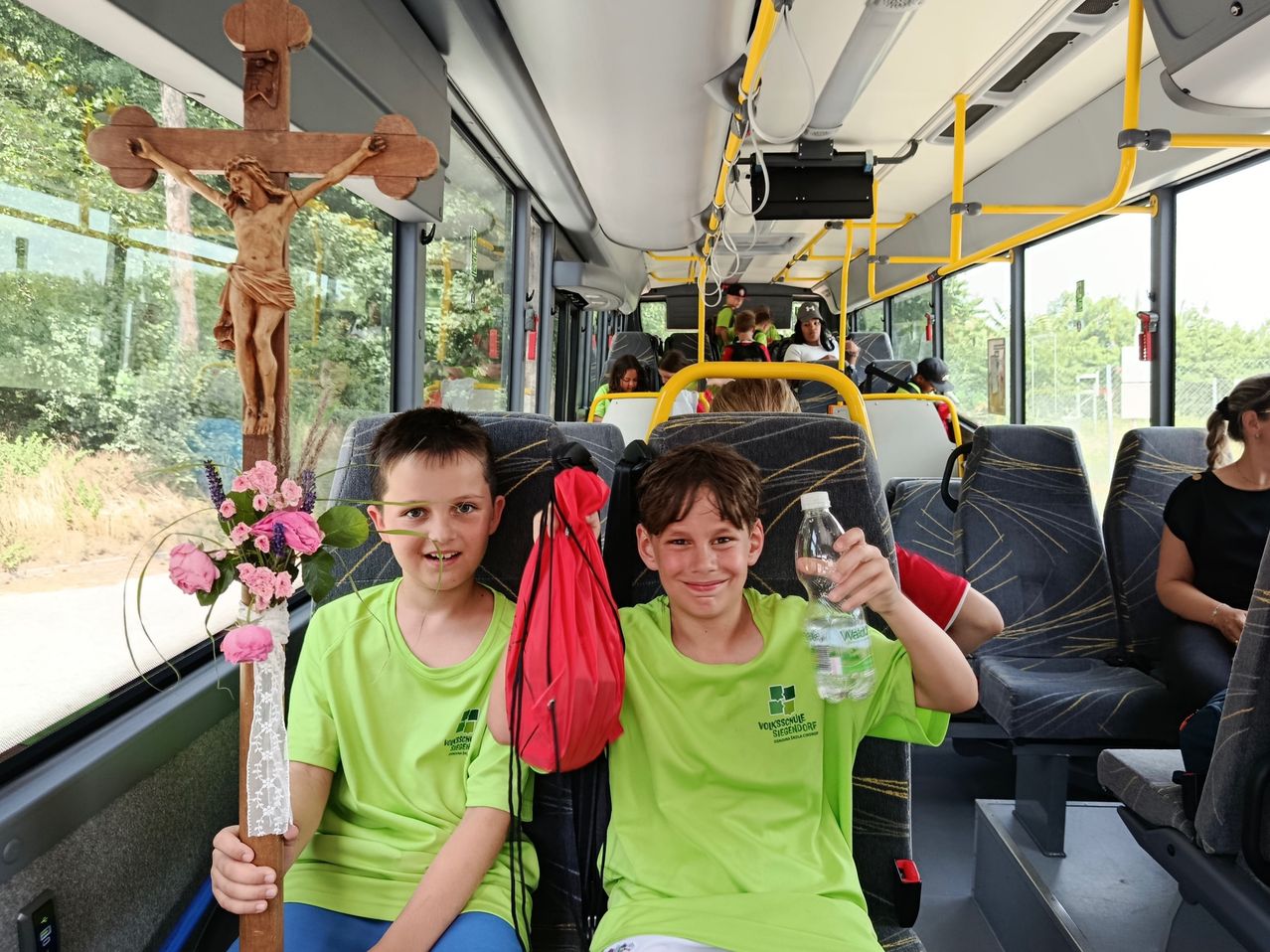 Zwei Jungen in grünen T-Shirts sitzen in einem Bus mit einem Kreuz und Blumen. Andere Passagiere sitzen hinter ihnen. Bäume und Gebäude sind durch die Fenster sichtbar.