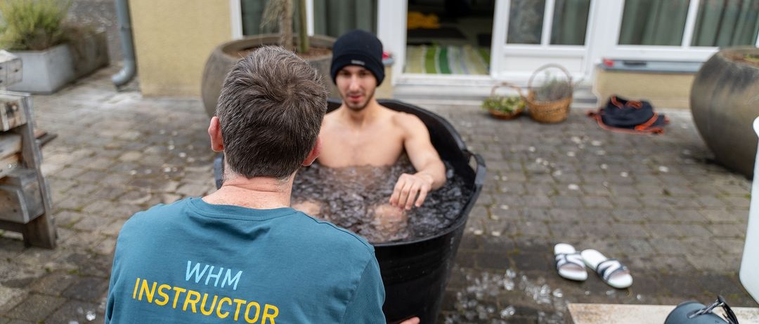 Ein Mann sitzt in einem schwarzen Eimer, der mit Eiswasser gefüllt ist. Ein anderer Mann mit einem blauen Shirt, auf dem 'WHM INSTRUCTOR' steht, sitzt neben ihm. Ein Gebäude mit Glasfenstern steht hinter ihnen.