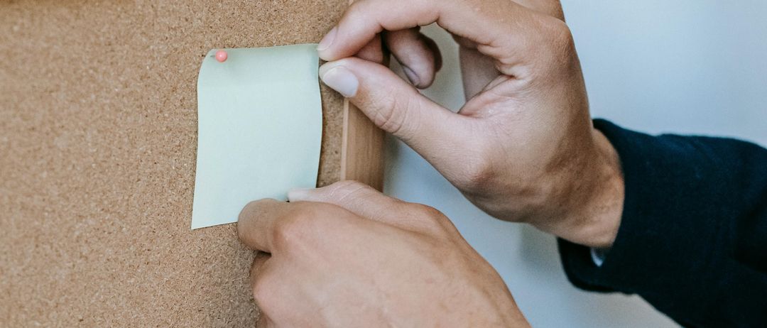 Eine Person heftet einen hellgrünen Haftzettel an eine Pinnwand, auf der bereits verschiedene farbige Notizen angeheftet sind.