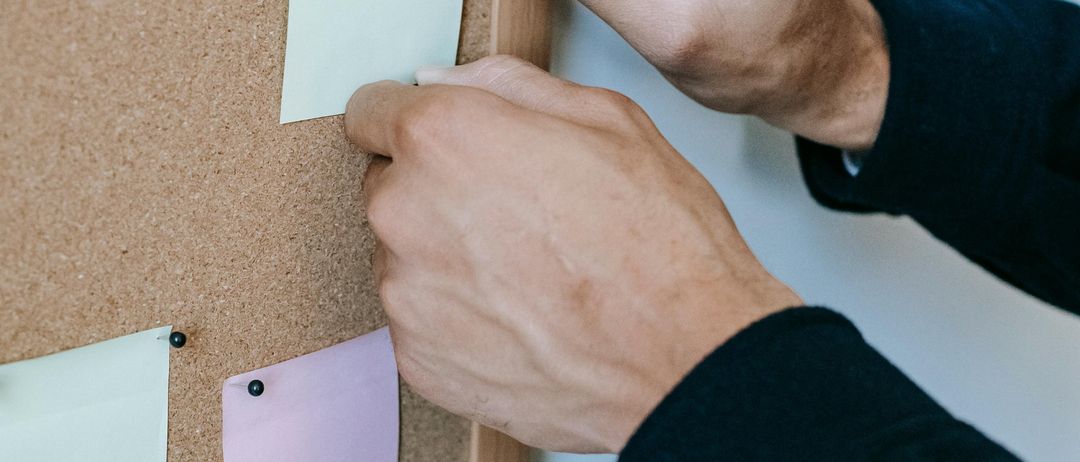 Eine Person heftet eine Haftnotiz an eine Pinnwand. Die Tafel ist braun und hat bereits mehrere Notizen. Die Person trägt ein dunkles Langarmshirt.