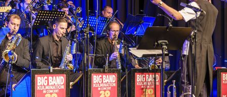 Ein Jazz-Orchester spielt auf der Bühne mit Saxophonen, Trompeten und einem Dirigenten. Die Bühne ist mit roten Schildern dekoriert, die 'Big Band Kulmination im Konzert 20 Jahre BBK' lesen.