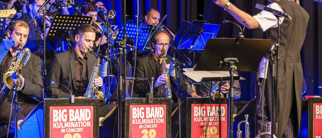 Ein Jazz-Orchester spielt auf der Bühne mit Saxophonen, Trompeten und einem Dirigenten. Die Bühne ist mit roten Schildern dekoriert, die 'Big Band Kulmination im Konzert 20 Jahre BBK' lesen.