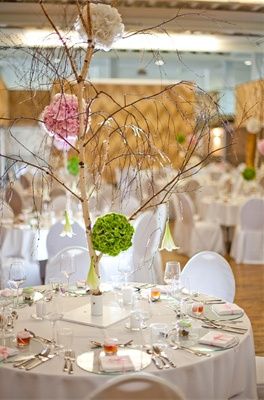 Ein Tisch für eine Veranstaltung mit einem Gesteck aus einem Baumzweig mit rosa und grünen Blumen, umgeben von weißen Stühlen und Tischen. Der Raum ist gut beleuchtet mit natürlichem Licht.