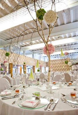 Eine Hochzeitsfeier mit einem runden Tisch, der mit Tellern, Besteck und Gläsern gedeckt ist. Der Tisch ist mit dekorativen Blumen-Zentren geschmückt und von Stühlen umgeben. Der Raum hat weiße Wände und Decken mit dekorativen Lichtern.