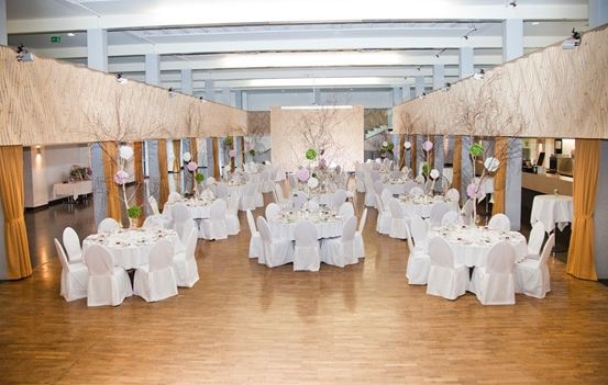 Ein Bankettsaal mit weißen Tischen und Stühlen, Holzböden und dekorativen Elementen wie Zweigen mit Blumen und Luftballons.