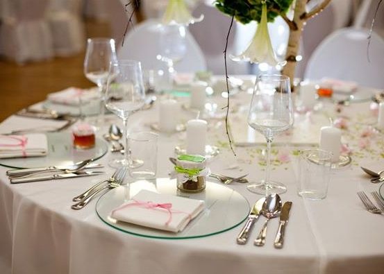 Ein Tisch ist gedeckt mit Tellern, Weingläsern, Besteck und Servietten. Ein kleines Glas mit einem grünen Band und eine Kerze ist auf jedem Teller platziert.