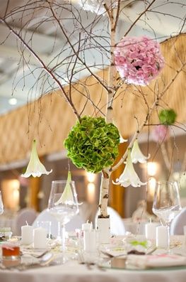 Ein Esstisch mit Wein glasses, Kerzen und einem Baum-Zentrenstück, das mit rosa, grünen und weißen Blumen dekoriert ist. Der Baum hat Zweige mit hängenden Blumen und steht auf einer weißen Tischdecke. Der Hintergrund ist unscharf.