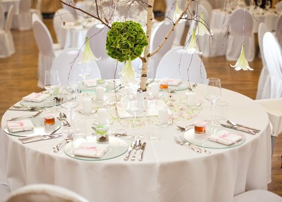 Ein runder Tisch ist mit weißen Tischdecken, Besteck und Gläsern gedeckt. Es gibt eine Tischdekoration mit einem Baumzweig, einer grünen Blume und weißen Blumen. Die Stühle sind weiß und um den Tisch herum angeordnet.