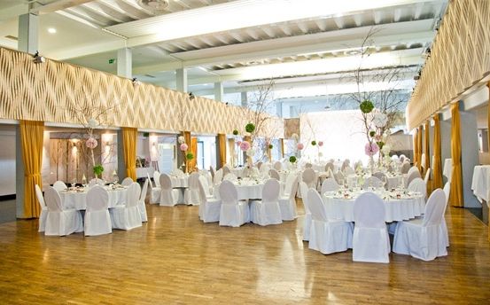 Ein großer Bankettsaal mit Holzboden und Tischen, die für eine Hochzeitsfeier vorbereitet sind. Die Tische sind mit weißen Tüchern bedeckt und haben Blumensträuße als Mittelpunkt.