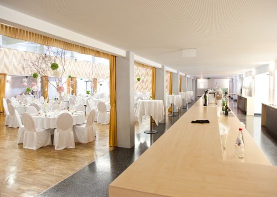 Ein Innenbankettsaal mit weißen Tischdecken und Stühlen. Der Saal hat einen langen Holztisch rechts mit Flaschen und Gläsern.