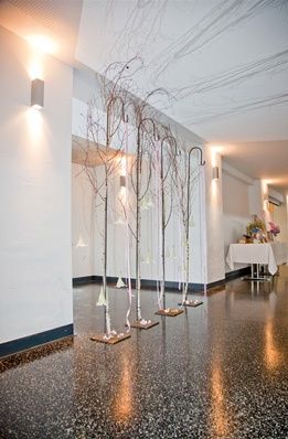 Ein leerer Raum mit einem glänzenden Boden, einem Tisch mit einer weißen Tischdecke und vier hohen weißen Bäumen mit nach oben ragenden Ästen.