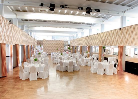 Ein großer Innenbereich, wahrscheinlich ein Bankettsaal, ist für eine besondere Veranstaltung dekoriert. Tische und Stühle sind in Reihen aufgestellt, mit weißen Bezügen und Blumenarrangements. Der Boden ist aus Holz, und die Wände haben ein Chevron-Muster.