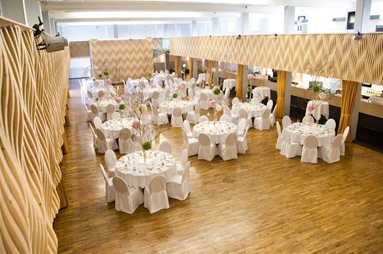 Ein großer Raum mit weißen Tischen und Stühlen, die in einem Rastermuster angeordnet sind. Jeder Tisch hat eine weiße Tischdecke, eine Vase mit Blumen und ein Tischmittelstück. Der Raum hat Holzböden und dekorative Wände.