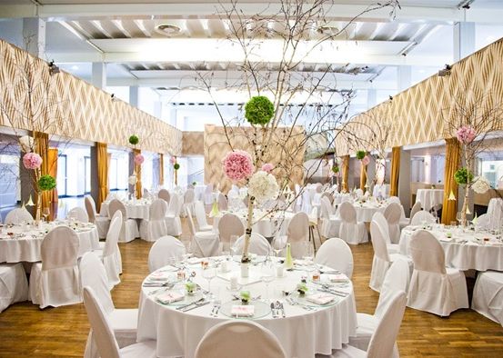 Ein großer Innenbereich für Veranstaltungen mit weißen Tischen und Stühlen. Die Tische sind mit weißen Tischdecken bedeckt und mit Blumenarrangements dekoriert. Der Raum hat Holzböden und eine hohe Decke.