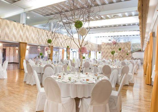 Eine Hochzeitsfeier im Innenbereich mit weißen Tischen, Stühlen und Blumendekorationen, Holzböden und orangefarbenen Vorhängen.