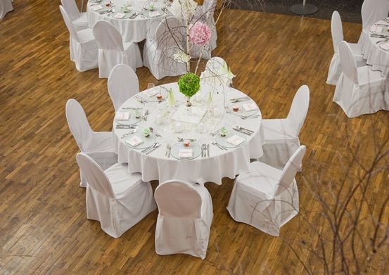 Ein Raum mit Holzböden, Tischen, die mit weißen Tischdecken bedeckt sind, Stühlen, die um die Tische herum angeordnet sind, und einem Mittelpunkt auf einem Tisch.
