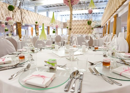 Ein großer Essbereich mit mehreren runden Tischen, die mit weißen Tischdecken, Servietten und Besteck gedeckt sind. Die Tische sind mit Blumen und Kerzen geschmückt.