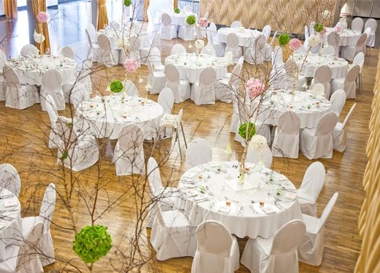 Ein Bankettsaal mit festlich gedeckten Tischen, geschmückt mit Blumen und Zweigen. Der Raum ist mit Stühlen gefüllt und hat Holzboden.