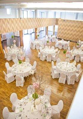 Ein Innenraum für Veranstaltungen mit mehreren runden Tischen, die mit weißer Tischwäsche bedeckt sind, eingerichtet für eine Hochzeit mit Blumenarrangements auf jedem Tisch, umgeben von weißen Stühlen. Der Raum hat Holzböden und hohe Decken.
