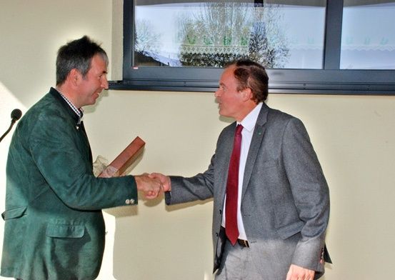 Zwei Männer in Anzügen und Krawatten schütteln sich die Hände in einem Raum mit einem Fenster und einem Mikrofon.