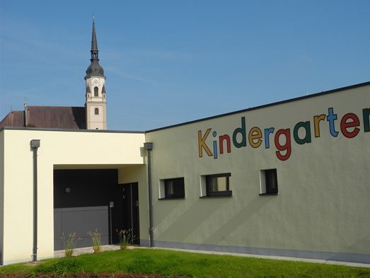 Die Vorderseite eines Kindergartengebäudes mit dem Wort Kindergarten in bunten Buchstaben an der Wand. Im Hintergrund ist ein Kirchturm zu sehen.