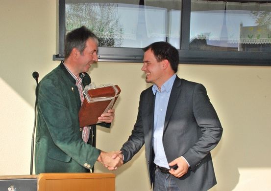 Zwei Männer schütteln sich in einem Raum die Hände, einer hält ein verpacktes Geschenk. Hinter ihnen steht ein Podest und ein Fenster, das Bäume zeigt.