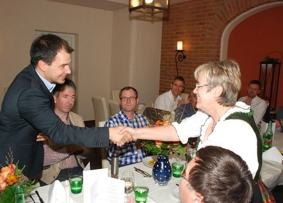 Ein Mann und eine Frau schütteln sich die Hände bei einem Treffen. Sie sind von Menschen umgeben, die an einem Tisch sitzen, mit Blumen und Tellern darauf.