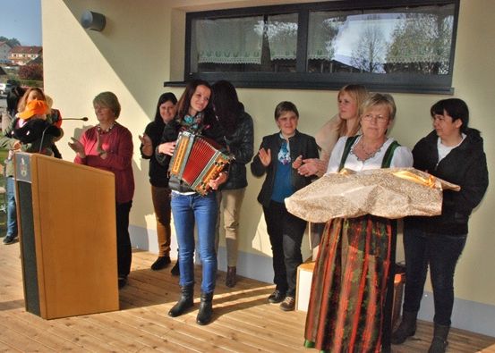 Eine Gruppe von Menschen, einschließlich Frauen und Kinder, steht auf einer Holzterrasse vor einem Gebäude und hält Musikinstrumente. Eine Frau spielt Akkordeon, während eine andere Frau ein eingewickeltes Objekt hält.